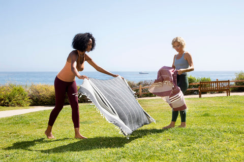 Two women airing a large flat-woven towel in the sun on a clifftop lawn