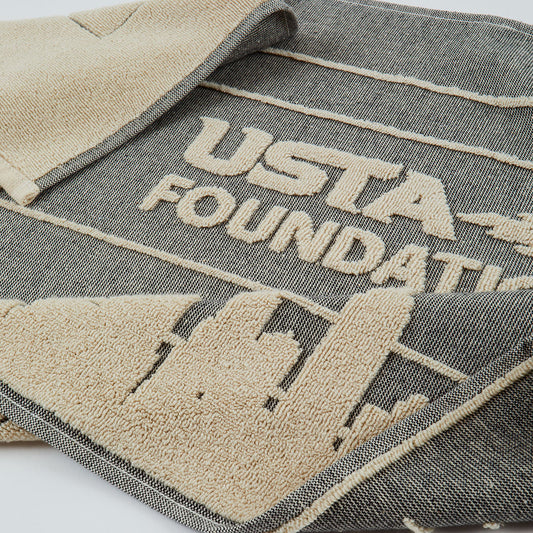 A close-up of a dark gray woven towel with beige embroidery featuring the text 'USTA FOUNDATION' and a building silhouette.