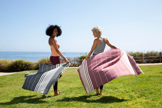 Two women holding open large flat-woven towels on a lawn by the sea