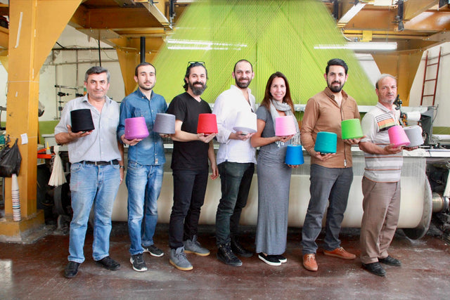 The team and mill staff standing in front of a loom, each holding a cone of coloured yarn
