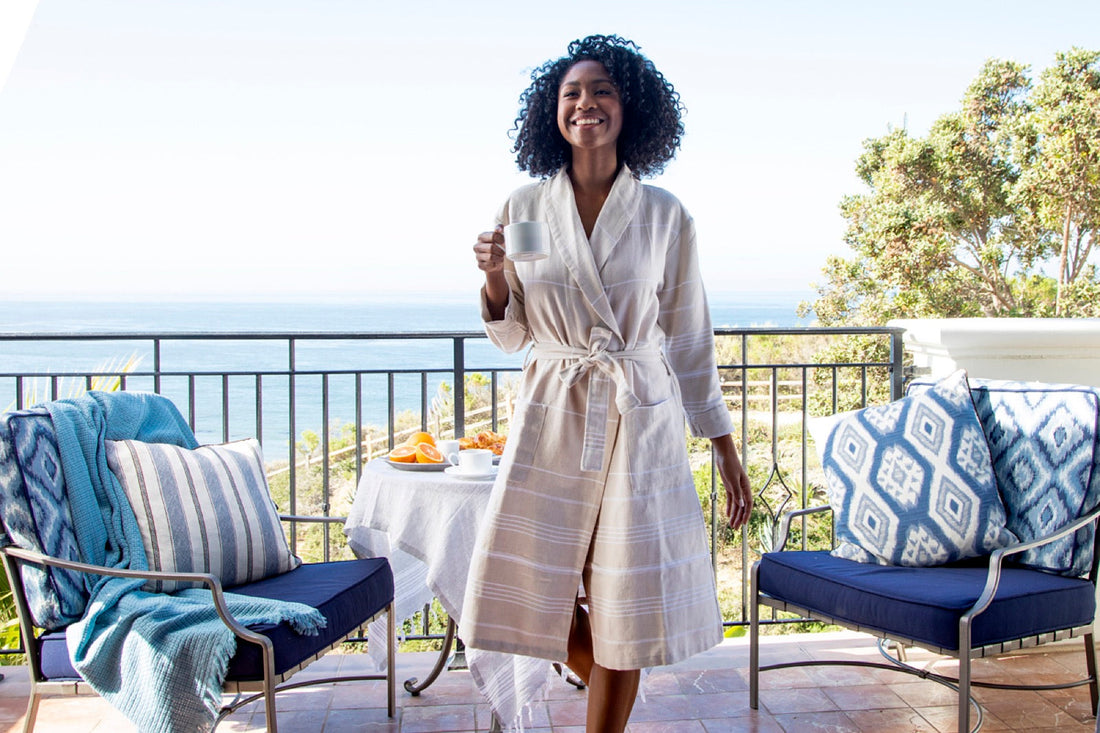 A woman in a beige robe holding a cup on a sunny balcony overlooking the sea