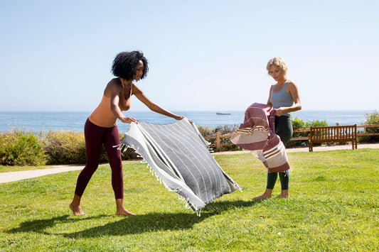 Two women airing a large flat-woven towel in the sun on a clifftop lawn