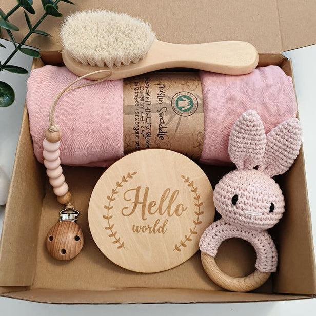 Baby care product set with pink blanket, wooden brush, teether, and rabbit-shaped ring in a cardboard box.