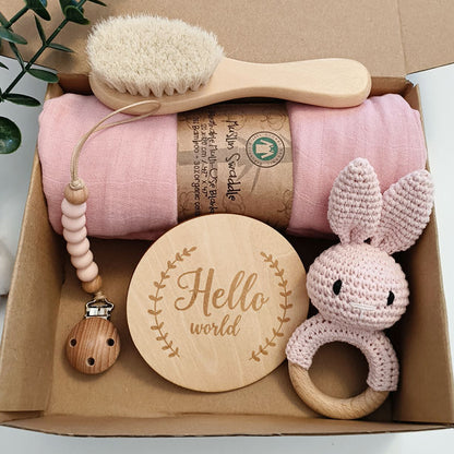 Baby care product set with pink blanket, wooden brush, teether, and rabbit-shaped ring in a cardboard box.