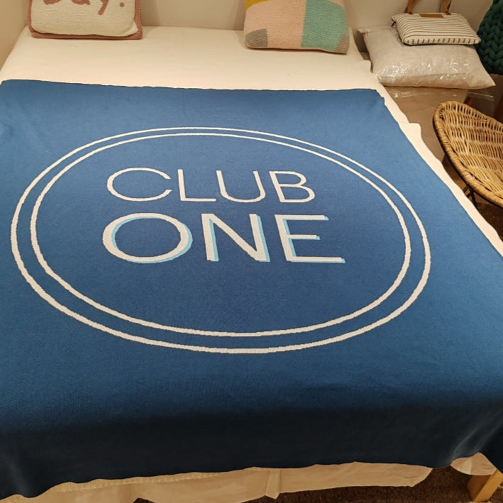 Blue blanket with 'CLUB ONE' text on a bed in a room.
