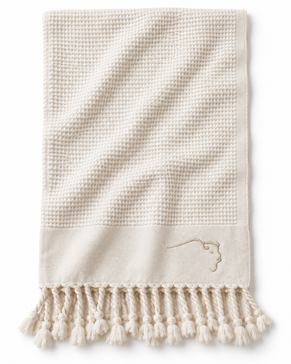 An off-white waffle-weave towel with a solid border, decorative fringe tassels, and a small embroidered logo on the lower right, shown against a plain white background.