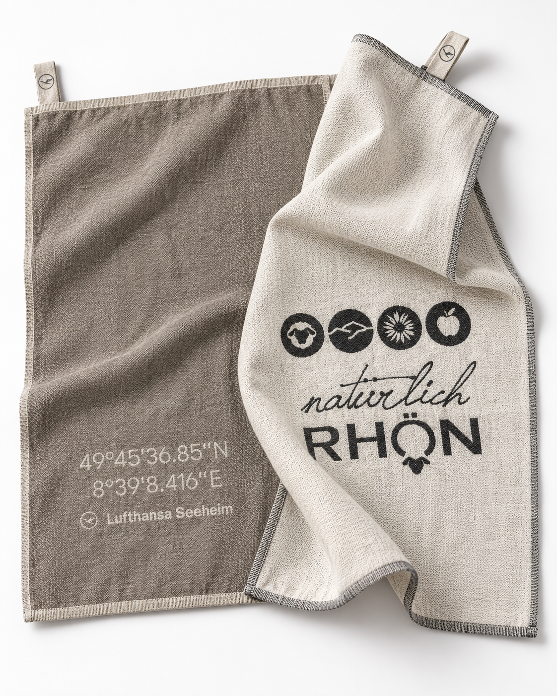 Two natural linen kitchen towels with gray trim, clipped at the top corners. One towel is taupe-brown with white printed coordinates and 'Lufthansa Seeheim' text, while the other is off-white with black printed icons and the words 'natürlich RHÖN'.