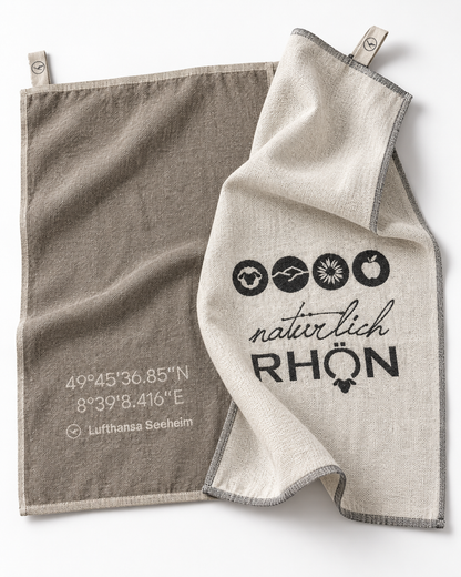Two natural linen kitchen towels with gray trim, clipped at the top corners. One towel is taupe-brown with white printed coordinates and 'Lufthansa Seeheim' text, while the other is off-white with black printed icons and the words 'natürlich RHÖN'.