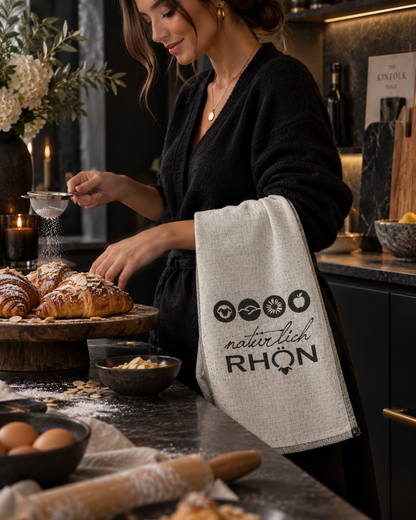 A woman in a black robe stands in a kitchen, sifting flour over croissants. A light beige linen towel with the black text 'naturally RHON' hangs off her arm, with baking props like eggs, a rolling pin, and bowls on the counter.