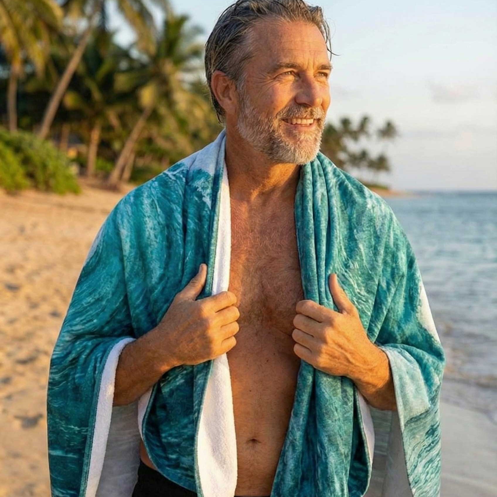 Man standing on a beach with a towel draped over his shoulders, surrounded by palm trees and ocean.