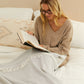 Woman reading a book on a bed with a herringbone grey and white blanket and pillow in the background