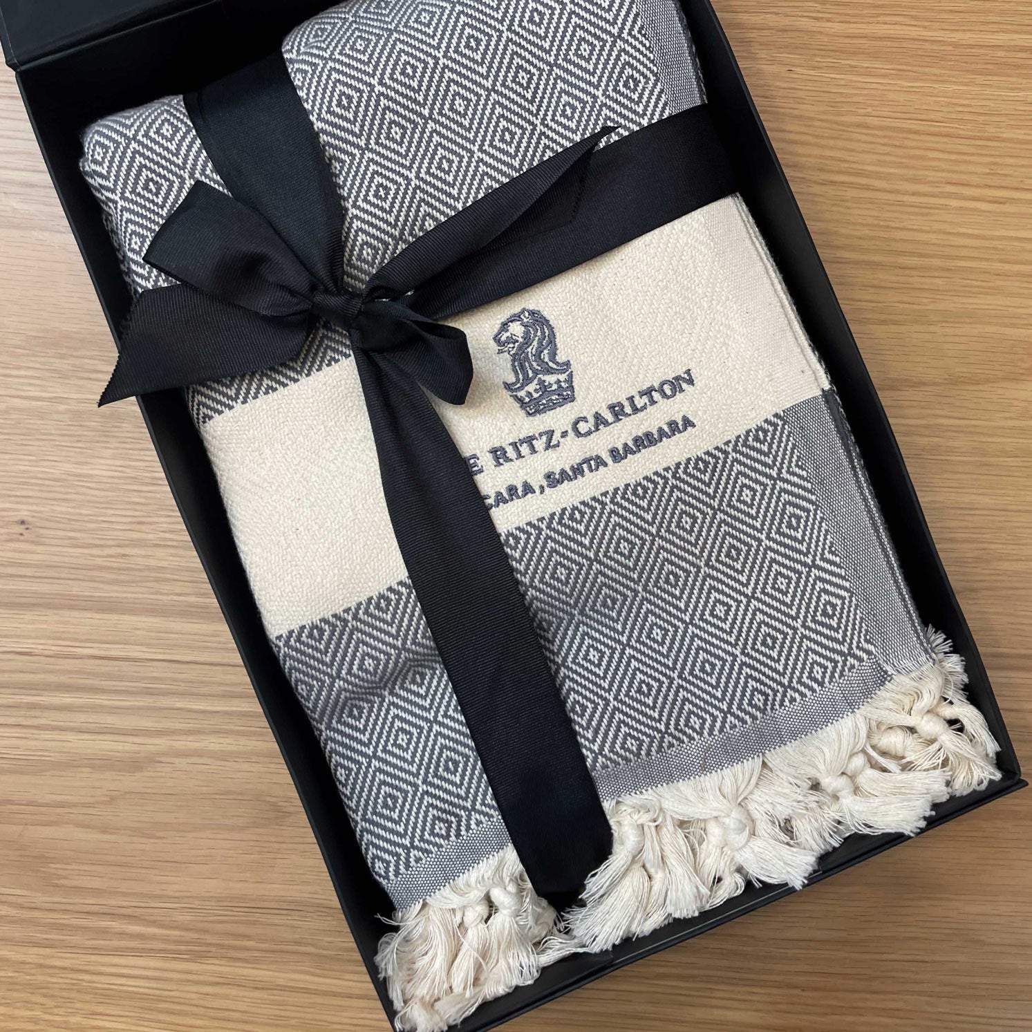 Folded blanket with geometric diamond pattern and black bow in a black box and embroidery of the Ritz Carlton Logo in grey embroidery yarns.