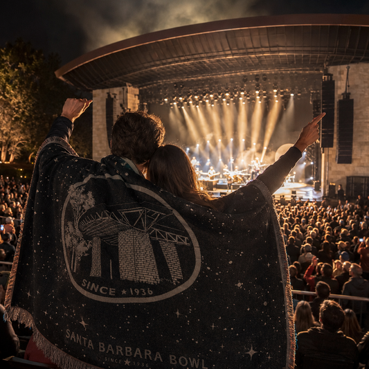 A crowd at an outdoor concert at night, with a person in the foreground holding up a dark Santa Barbara Bowl themed blanket featuring a large white bowl logo and the text 'SANTA BARBARA BOWL SINCE 1936'.