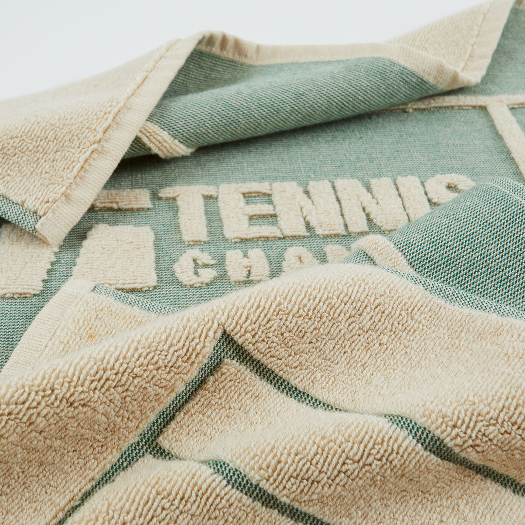 A close-up of a woven towel or blanket with a sage green and beige color-blocked design. The beige side features a plush terry texture, while the green side shows a woven pattern with the words 'TENNIS CHAMP' stitched into it.
