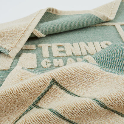 A close-up of a woven towel or blanket with a sage green and beige color-blocked design. The beige side features a plush terry texture, while the green side shows a woven pattern with the words 'TENNIS CHAMP' stitched into it.