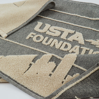 A close-up of a dark gray woven towel with beige embroidery featuring the text 'USTA FOUNDATION' and a building silhouette.