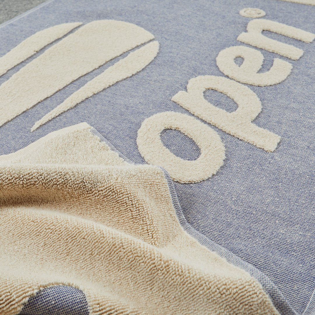 Close-up of a navy and beige textured towel or blanket with large raised letters spelling 'open' and tennis ball shapes, draped over a blue-gray woven surface.