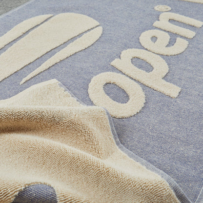 Close-up of a navy and beige textured towel or blanket with large raised letters spelling 'open' and tennis ball shapes, draped over a blue-gray woven surface.