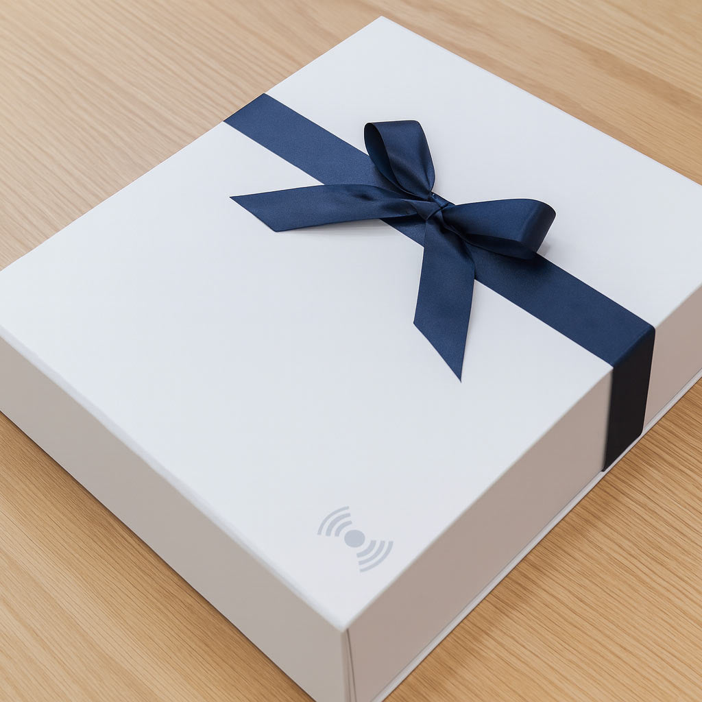 White box with a blue ribbon on a wooden surface. The icon on the box indicates that it has a magnetic closure