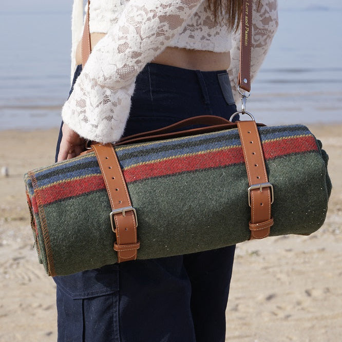 Person holding a rolled-up green picnic blanket with red and blue stripes and a brown leather strap.
